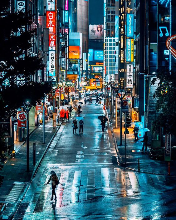 a view looking down a rainslicked street in a japanese city. people with umbrellas walk down the street, which is lined with brightly lit signs. the reflection of the lights creates a colorful, vibrant atmosphere.