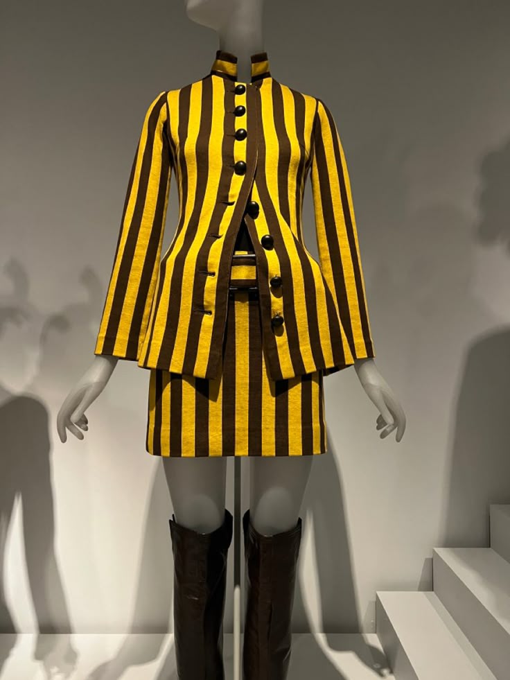 a mannequin dressed in a yellow and brown striped skirt and jacket ensemble with brown boots, part of a fashion exhibit. the jacket has a mandarin collar and large black buttons. the background is a minimalist exhibit setup with stairs.