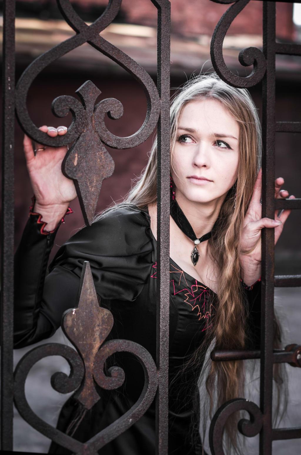 a medium shot captures a young woman with long blonde hair, wearing a black dress with red trim and a black choker, looking away while standing behind an ornate wrought iron gate. her hands are lightly resting on the gate as she gazes thoughtfully into the distance.