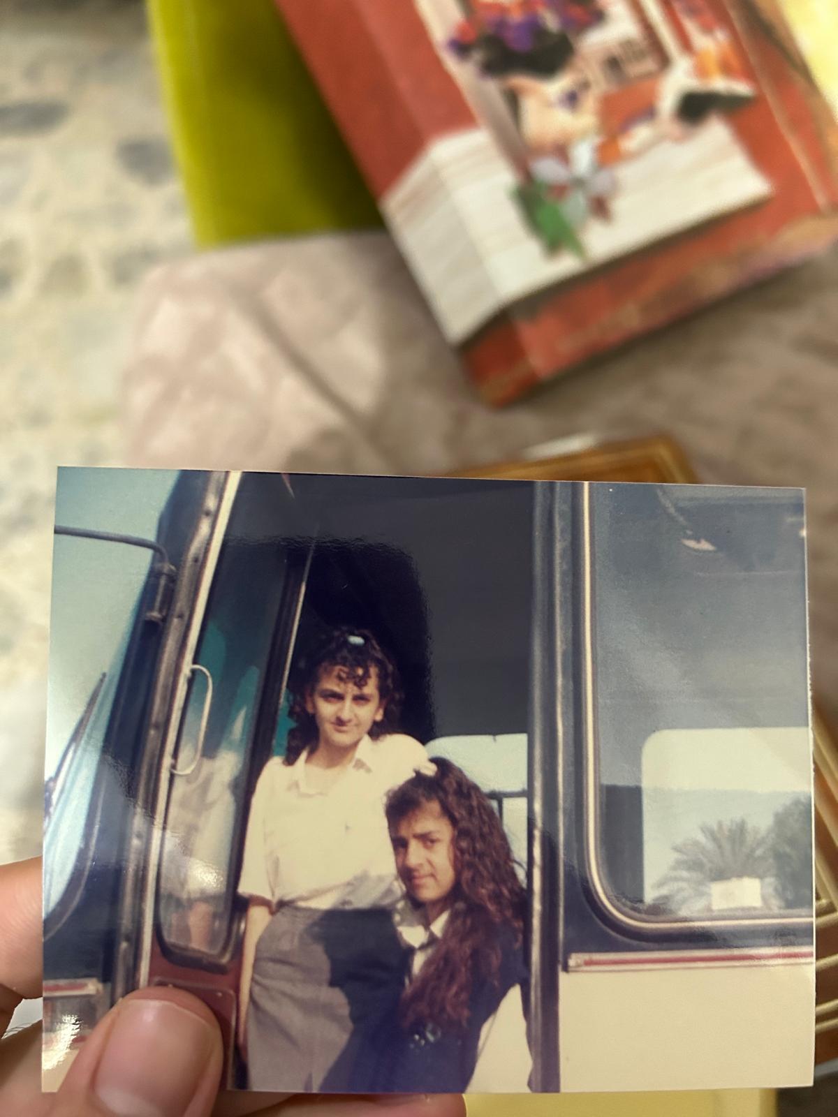 the image is a closeup photograph of a vintage print showing two young women sitting in the doorway of a bus.  the photo appears to be from the 1980s or early 1990s, judging by the clothing styles and overall feel. one woman has shoulderlength curly brown hair and is wearing a lightcolored, possibly white, longsleeved shirt and a darker skirt or pants.  the other woman has long, curly dark brown hair and is wearing a darkcolored jacket or top.  they appear to be friends, and both have smiles on their faces. the background shows the interior and exterior of the bus. the photo itself appears slightly faded or aged, further enhancing its vintage appearance. the photograph is held by a hand in the bottom left corner.
