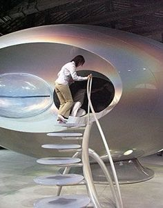 two women are entering a large, metallic, futuristic spaceshiplike structure through an open hatch. the spaceship has a shiny, iridescent exterior and a clear, domeshaped window. a curved staircase with translucent steps leads up to the hatch. the environment appears to be an indoor setting, possibly a museum or exhibition hall, with bright, natural lighting.