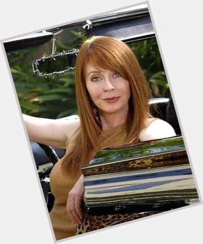 a woman with long red hair is wearing a brown jacket and looking directly at the camera while standing near a car.