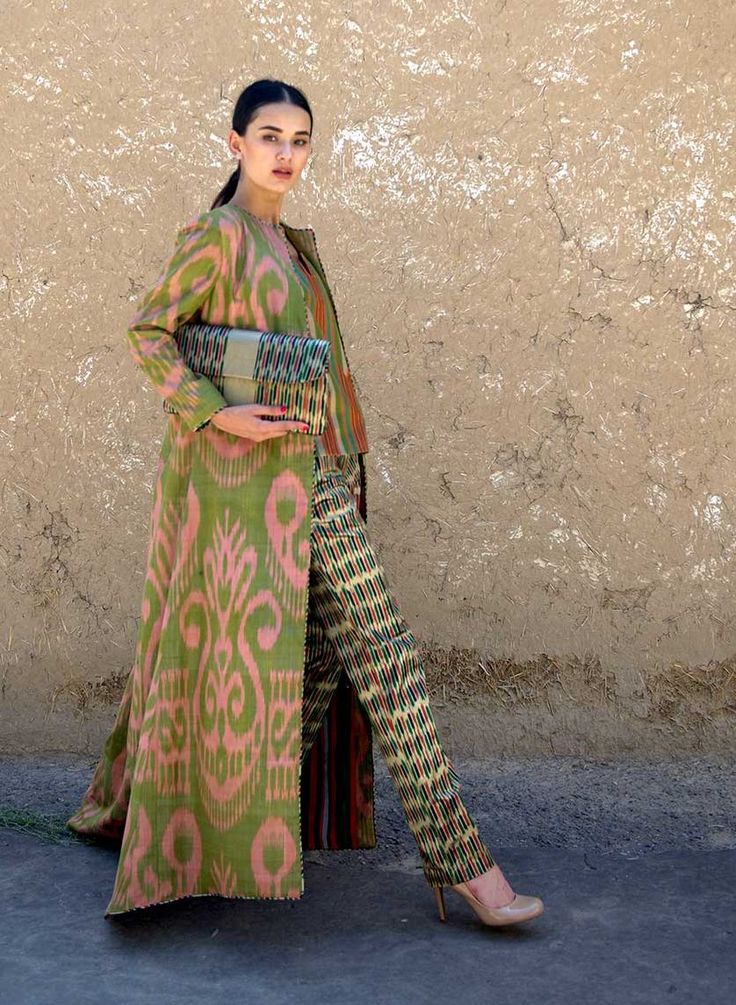 a stylish woman is standing against a rustic earthen wall. she's wearing a long, flowing coat and matching pants, both made of vibrant ikat fabric.  the ikat pattern is predominantly green, pink, and orange with intricate designs. she carries a matching clutch. the outfit is elegant and sophisticated, blending traditional central asian designs with modern fashion sensibilities. the woman's pose and overall look suggest a fashion shoot or editorial photo.  the background highlights the texture of the wall, providing a contrast to the smooth textures of the fabrics.