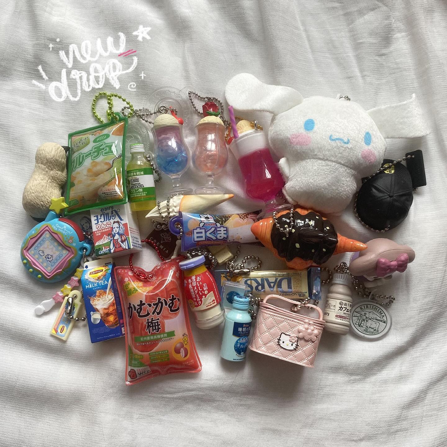 a collection of kawaii charms and keychains, predominantly food and drink themed, arranged on a white surface. many of the items are miniatures of japanese snacks and beverages. there are also charms of sanrio characters like hello kitty and cinnamoroll. additionally, there are small hat, purse, and tamagotchi charms.