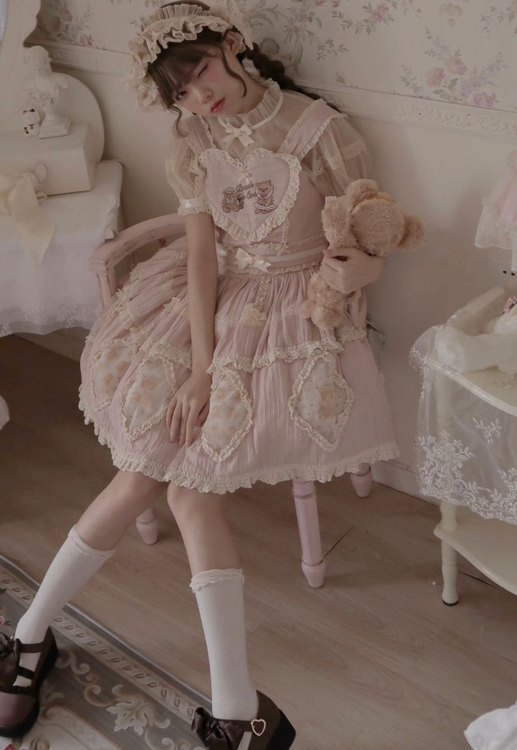 a young woman is posing in a pink lolita fashion dress. she is wearing a matching headband and holding a teddy bear. the dress is adorned with lace, ruffles, and floral details. the room is decorated in a vintage style with floral wallpaper and white furniture.