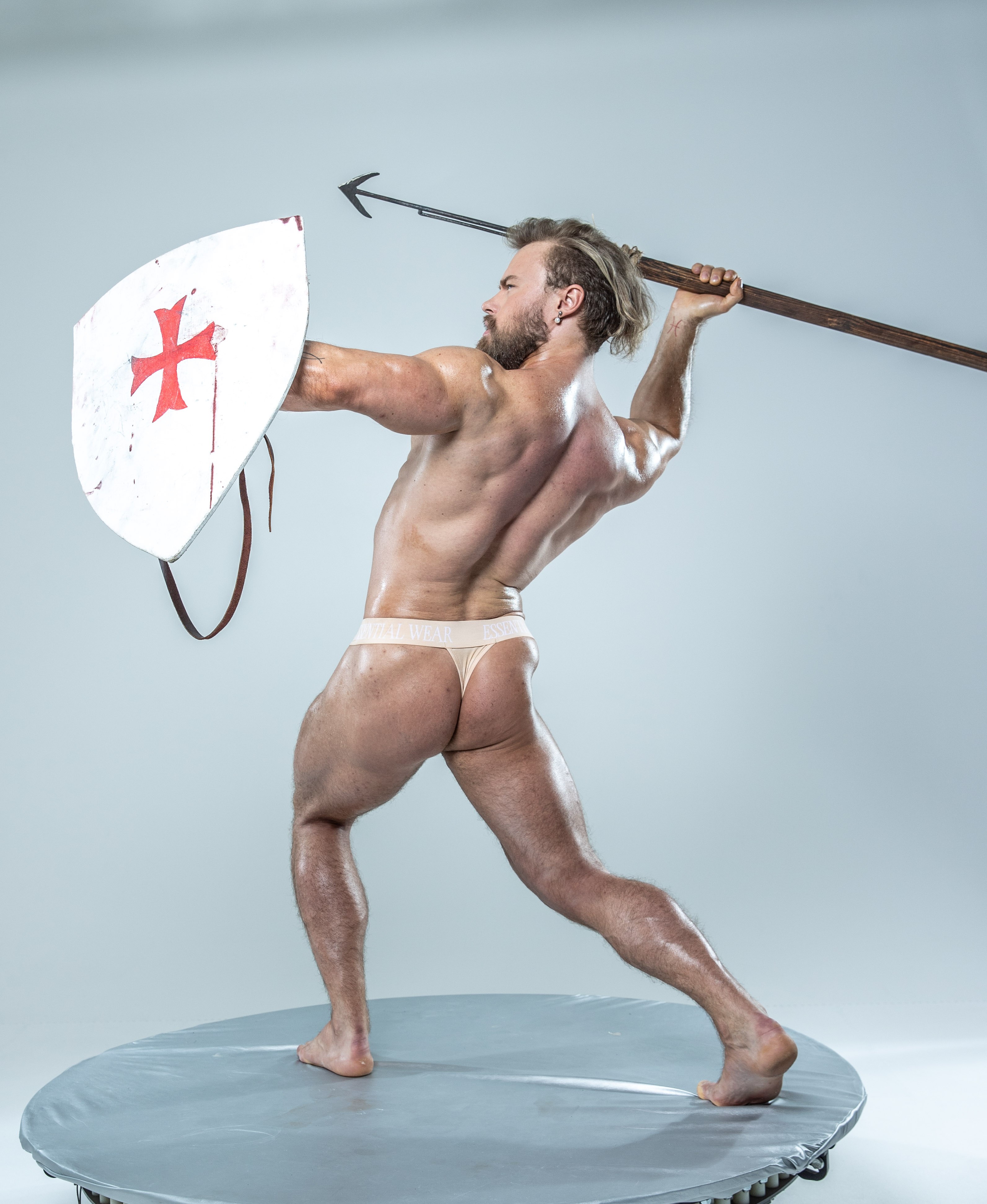 a muscular man in minimalist underwear stands in a dynamic pose against a plain gray background. he holds aloft a spear in his right hand and a  round shield in his left, seemingly in midbattle. the shield features a red cross on a white field. the image conveys a sense of strength, power, and ancient warfare.  the studio lighting illuminates the scene brightly.