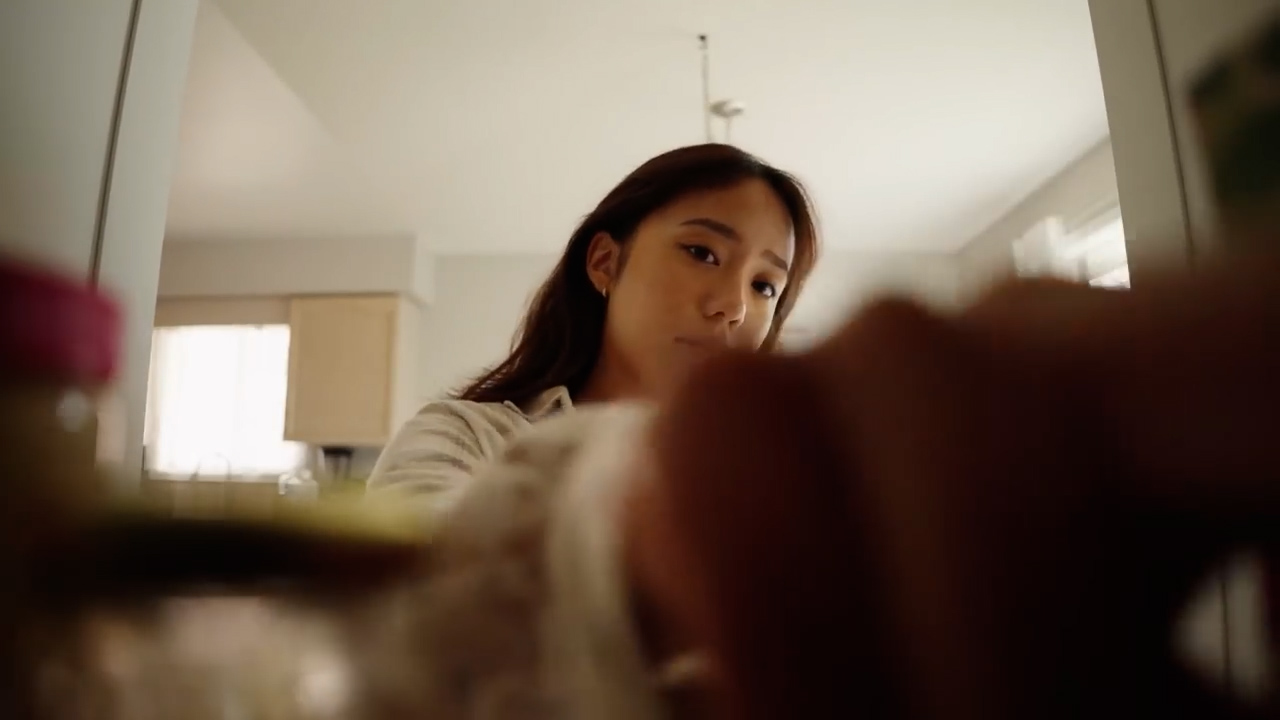 the image offers a closeup, lowangle perspective of a young woman in her kitchen.  she is slightly out of focus, suggesting movement, and seems to be reaching into a refrigerator or cabinet. the warm, natural lighting and the glimpse of food items hint at a scene of everyday domesticity, likely meal preparation. the low viewpoint adds a sense of intimacy and places the viewer directly in the scene.