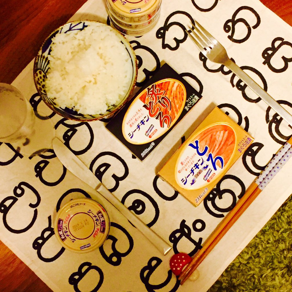 an overhead photo of a place setting for one person. there's a bowl of rice, four cans of tuna, a glass, a fork, a knife and chopsticks on the place mat. the place mat has a repeating pattern of a letter from the sinhala alphabet.