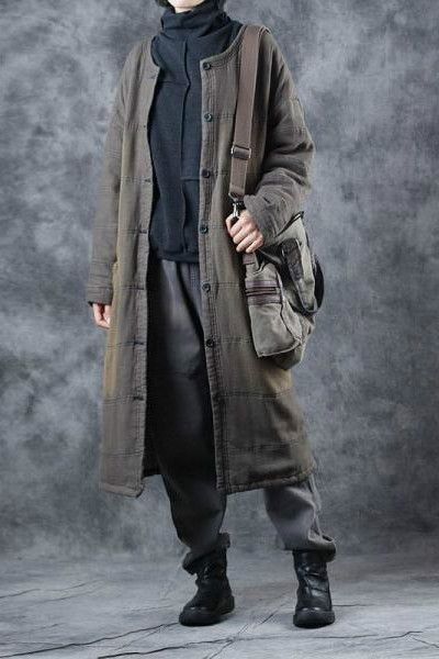 a woman is standing in a stylish long coat with a black turtleneck sweater. she is wearing grey trousers and black boots with a bag hanging on her shoulder.  she is looking down with a  straight face.