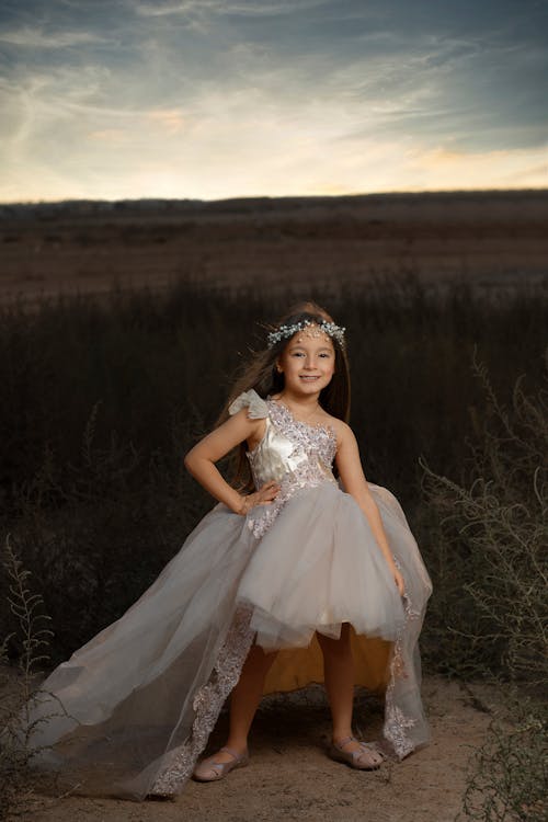 a young girl, possibly around 710 years old, is the focus of this outdoor portrait. she wears a stunning, light beige highlow tulle dress adorned with lace detailing. the dress has a oneshoulder design and features a long train in the back.  she's also wearing a delicate flower crown. the girl stands in a field of tall grass, with a sunset backdrop creating a warm, hazy atmosphere. her expression is happy and she appears confident and graceful. the overall scene is idyllic and picturesque, suitable for a magazine or fashionrelated publication.