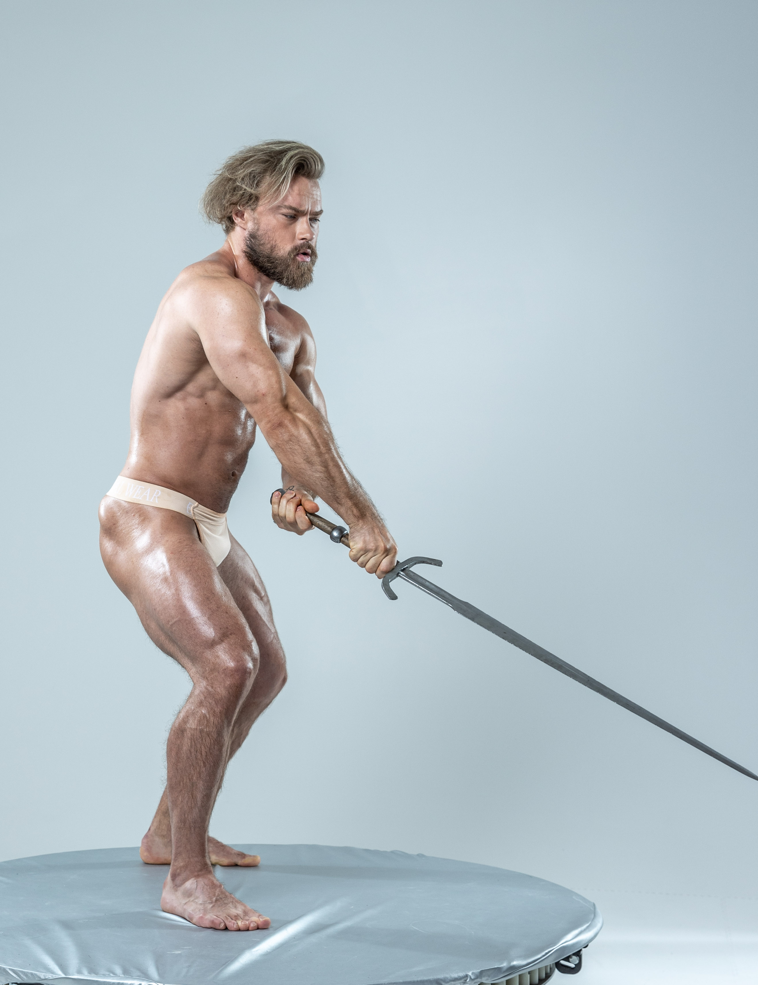 a muscular man with long blonde hair and beard, wearing only a beige loincloth, stands in an attack stance on a round gray platform against a light gray background. he grips a doubleedged sword with both hands, preparing to swing it.  the image is professionally lit, highlighting the man's physique and the intensity of the pose.