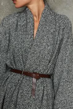 a close up shot of a woman wearing a black and white herringbone coat with a brown leather belt. she is looking away from the camera. the image is taken in a studio setting.