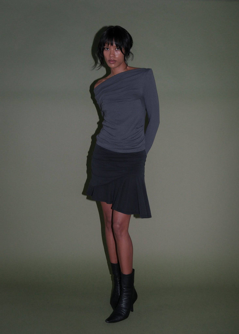 a fulllength studio shot of a woman wearing a grey oneshoulder longsleeved top and a short black skirt with ruffles, paired with black leather boots. she stands against an olive green background and has short dark hair.