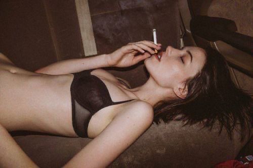 a woman in a black bra is lying down, smoking a cigarette. the image is a portrait shot indoors.