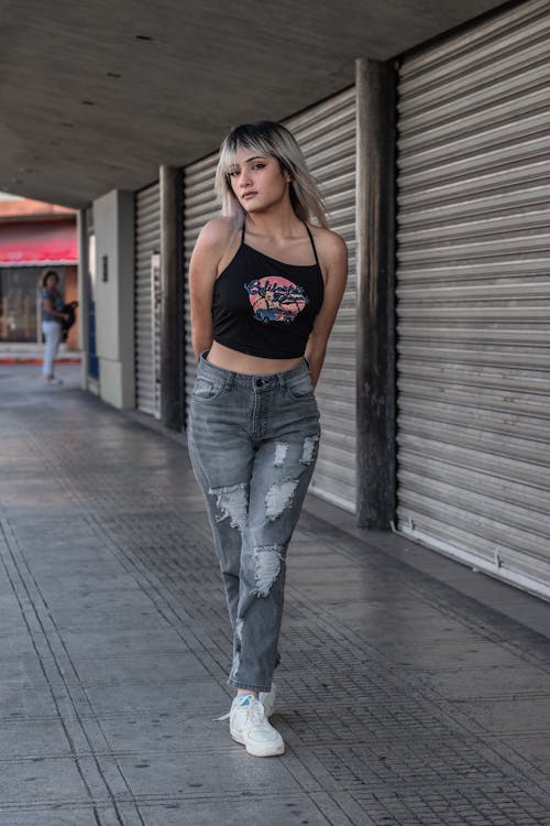 a young woman with blonde hair is standing on a city sidewalk, posing for a photo. she's wearing a black crop top, light gray ripped jeans, and white sneakers. the background includes a building with metal rolling shutters.  she appears confident and stylish.