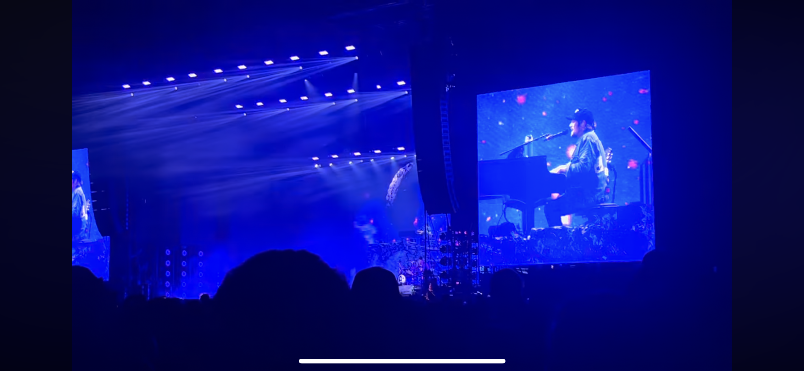 a concert venue filled with people, with the stage lit up in blue lights. a musician is performing on stage, playing the piano. the crowd is silhouetted in the dark, and the atmosphere is electric. it is a live music performance in a dark venue, with the stage brightly lit in blue lights. the audience is captivated by the performance.