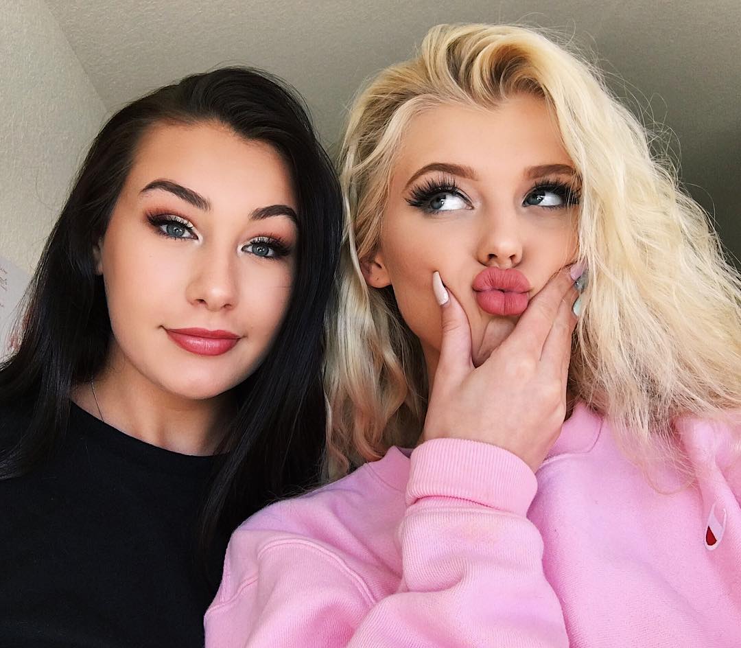 a selfie of two young women. one woman has long, dark hair and is wearing a black tshirt and red lipstick. the other woman has blonde, wavy hair and is wearing a light pink sweatshirt. she is holding her hand to her cheek and puckering her lips into a pout. both women have heavy makeup, including long eyelashes.