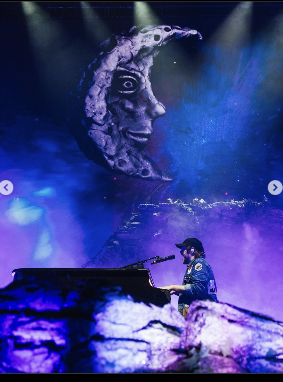 a musician is seen playing a piano on stage at a concert. he is silhouetted against a large image of a moon and a dark sky. the stage is brightly lit with spotlights. the performer is wearing a blue jean jacket and a black baseball cap.