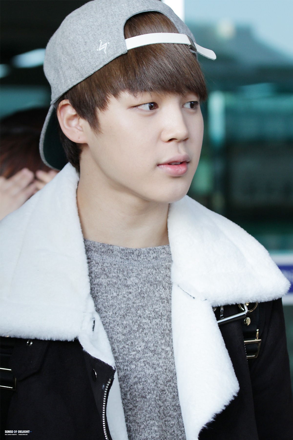 a portrait of park jimin wearing a grey cap, grey sweater, and a black coat with a white fur collar. he is looking off to the side.