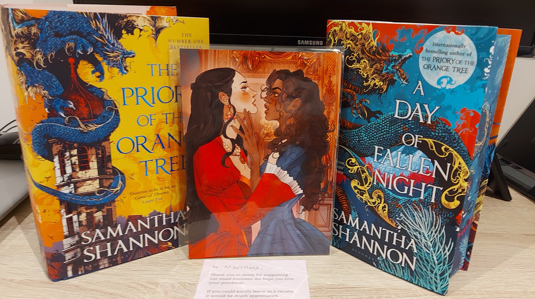 the image contains two books by samantha shannon, "the priory of the orange tree" and "a day of fallen night," along with an illustration featuring two women in period clothing. the book covers depict dragons. a small thank you note is visible in front of the illustration. the items are arranged on a desk.