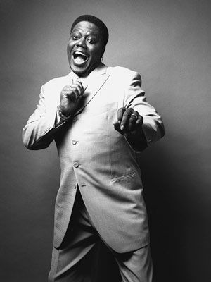 a black and white photo of a man in a suit. he is smiling and looking at the camera. he is wearing a white jacket with a white shirt underneath. the man is posed against a plain, white background.