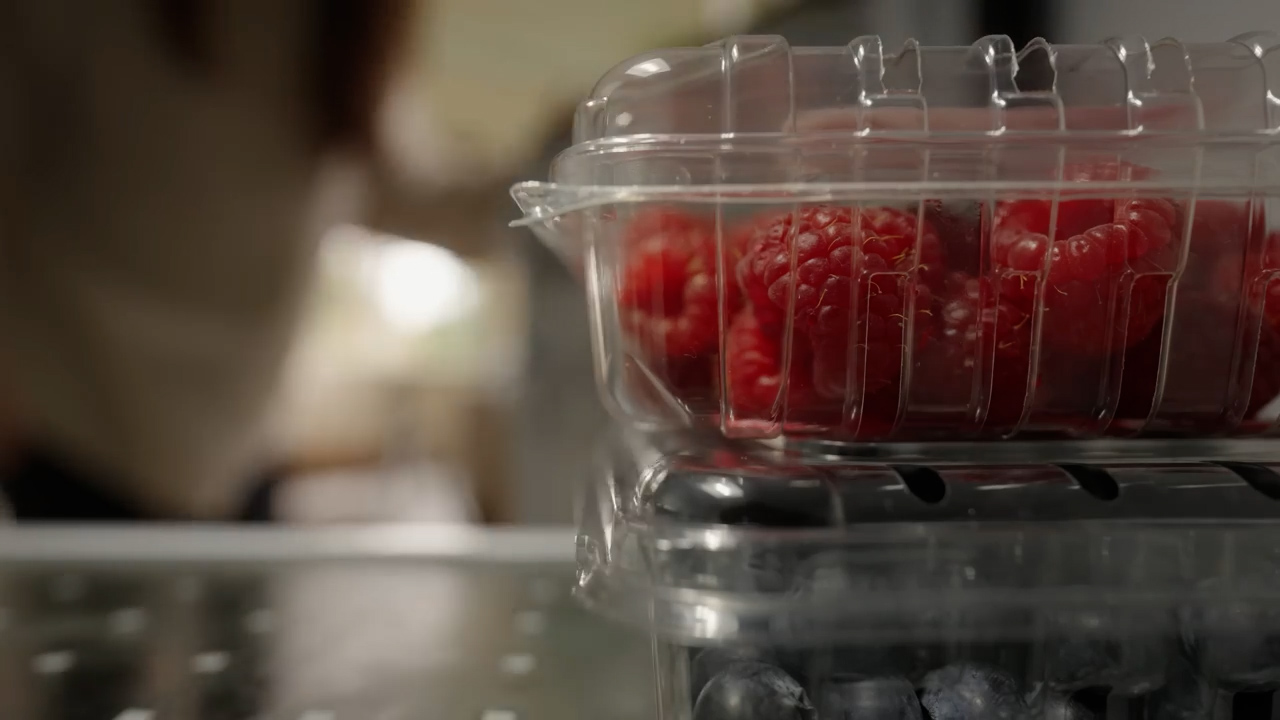the image shows a closeup view of a plastic container filled with fresh raspberries. the raspberries are stacked on top of another container likely filled with blueberries, partially visible at the very bottom of the frame. a woman, slightly out of focus, is visible in the background, suggesting she's in a kitchen setting.