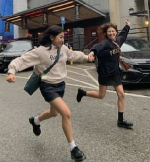 two girls are running in the street, one is wearing a white tshirt and black shorts and the other is wearing a black jacket and black shorts. both girls are smiling and having fun. there are cars parked in the background and there is an urban setting.