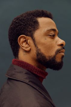 a man with a short afro fade haircut and a full beard. he is wearing a dark brown turtleneck sweater and a black jacket. he is facing to the right of the frame, and his eyes are looking straight ahead. he has a roman numeral "ii" tattoo behind his left ear. the background is a dark teal color, and the lighting is soft and diffused.
