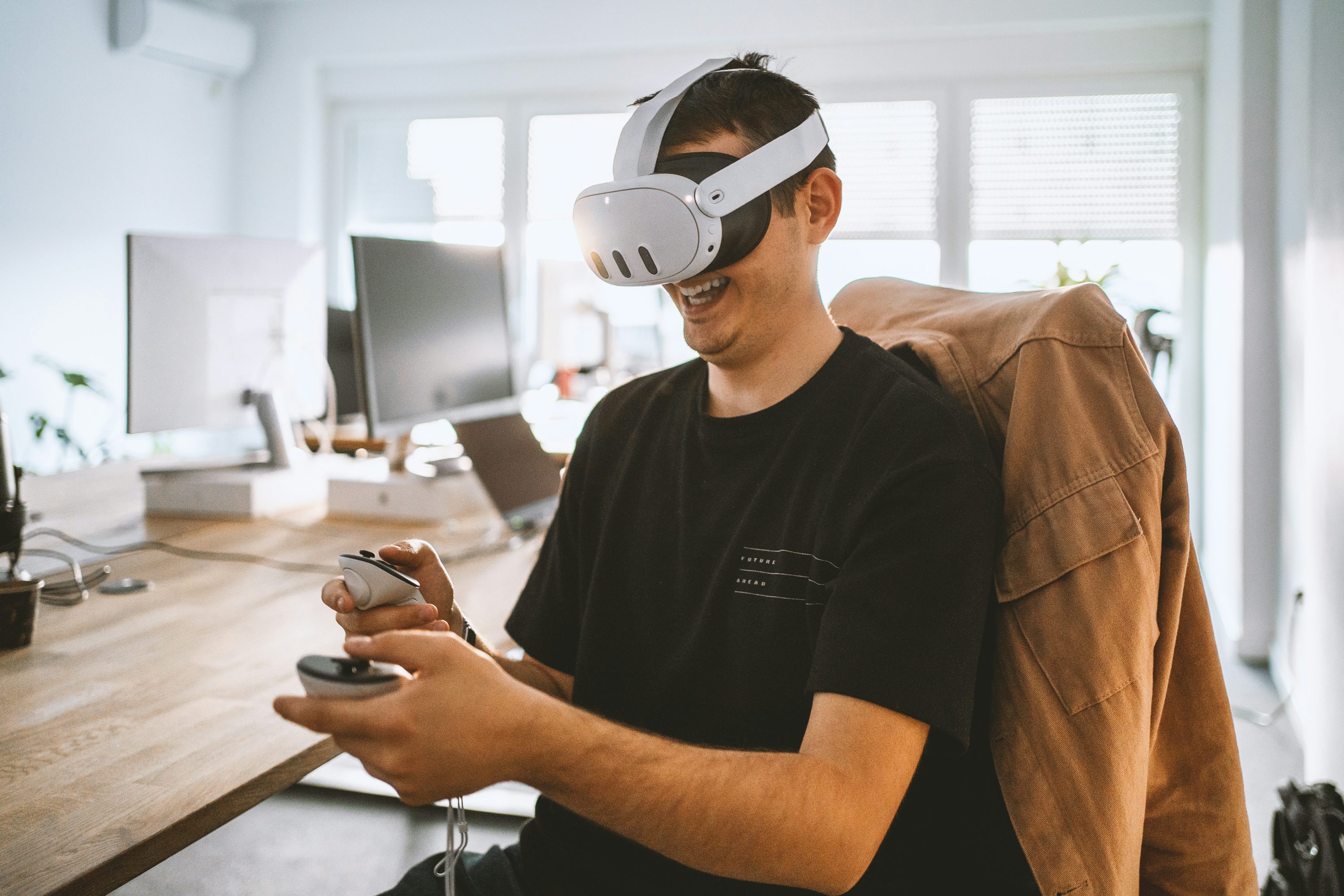 a man wearing a white vr headset is sitting at a wooden desk in a bright office. he is holding two white controllers and smiling, presumably playing a virtual reality game. there are two computer monitors and a laptop on the desk. a tan coat is draped over the back of his chair. a window with blinds provides natural light.