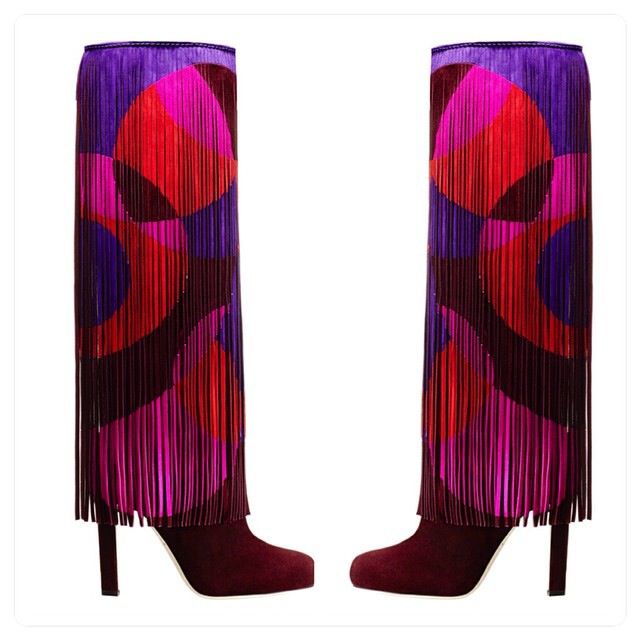 two kneehigh suede boots are displayed against a white background. the boots are a deep reddishpurple color at the foot and heel. above the foot, the boots feature long strands of fringe in various shades of purple, red, and pink arranged in a circular pattern. the heel of each boot is a slim, high heel in the same suede color as the foot. the boots are oriented with the toes facing each other.