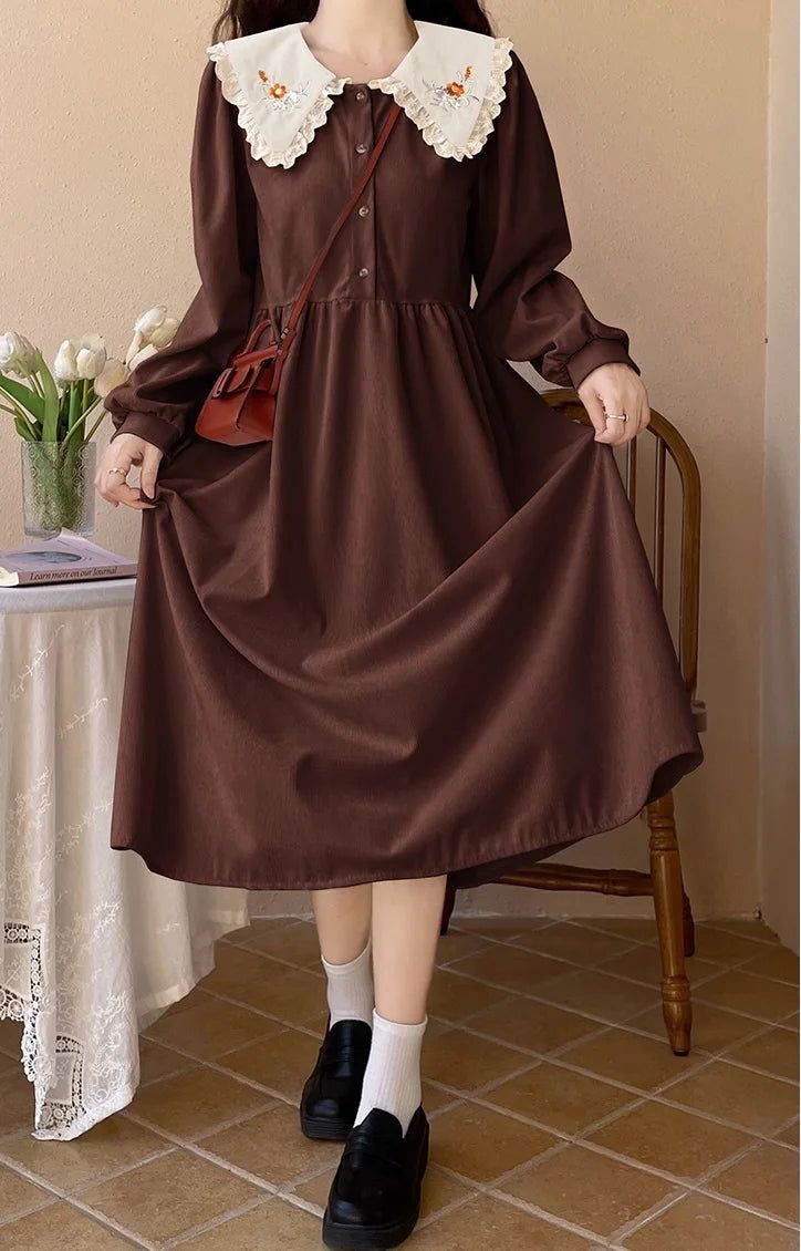 a woman is standing in a room, modeling a brown longsleeved midi dress with a large white collar embroidered with small flowers. she is wearing white socks and black loafers, and carrying a small brown purse.  the dress has a buttondown front and a flowing skirt. there is a table behind her with a vase of flowers and a book on it, and a wooden chair is visible to the right. the overall style of the image is whimsical and feminine, with a vintage or cottagecore aesthetic.