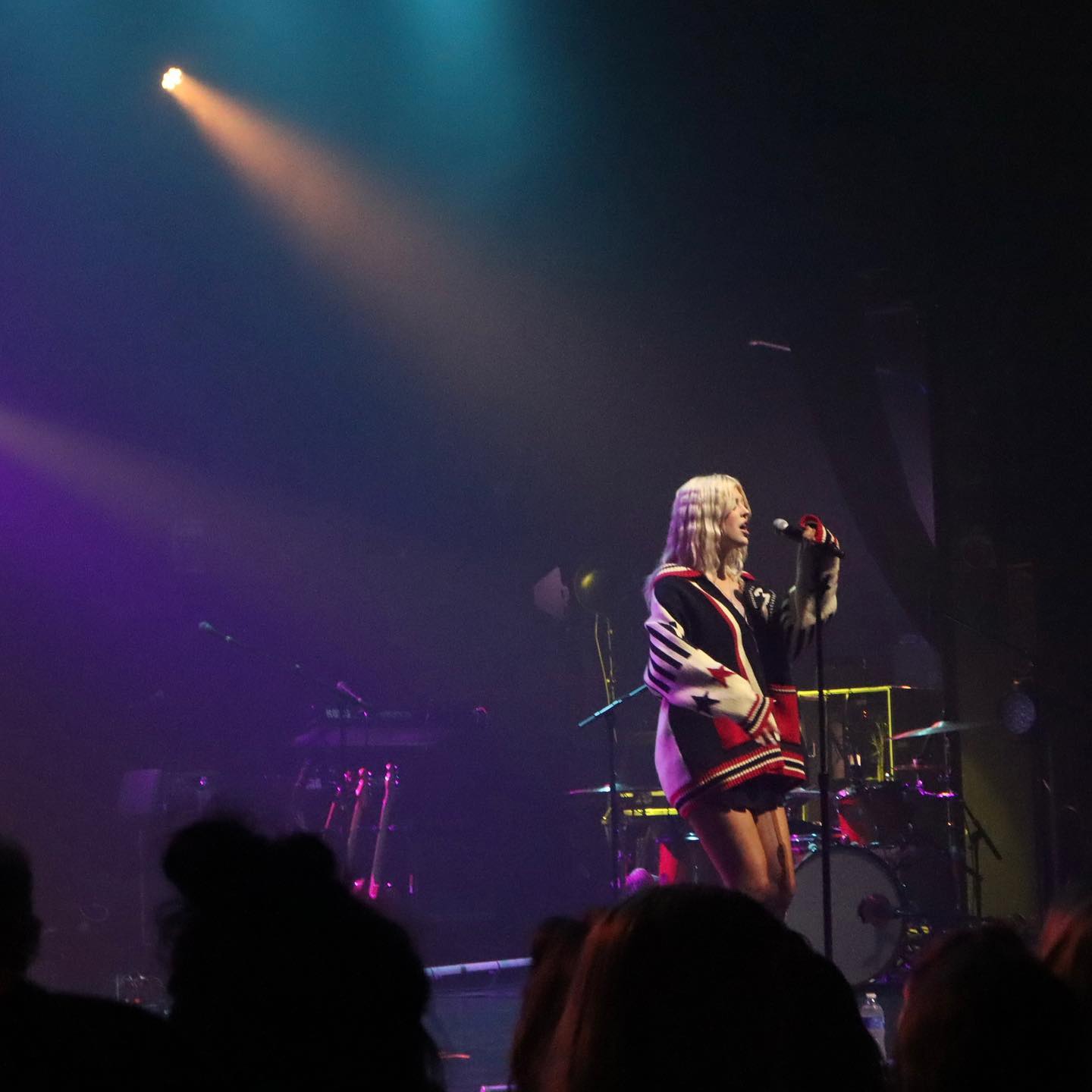 a woman with blonde hair wearing a jacket with red, white, and black designs is singing into a microphone on a stage during a concert. she is standing in front of a drum set and other musical equipment. the stage is lit with blue, purple, and orange lights. the audience is visible in the foreground.