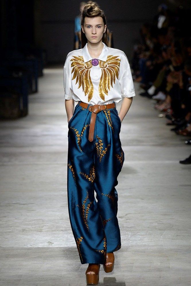 a runway model walks down the catwalk wearing a stylish outfit. the outfit consists of a white shirt with gold embellishments resembling wings, widelegged teal pants with gold detailing, and brown leather platform shoes. the model has her hair styled neatly and is displaying a confident demeanor. the background is blurred, showcasing other audience members and elements of the runway environment. the overall image conveys a sense of high fashion and elegance.
