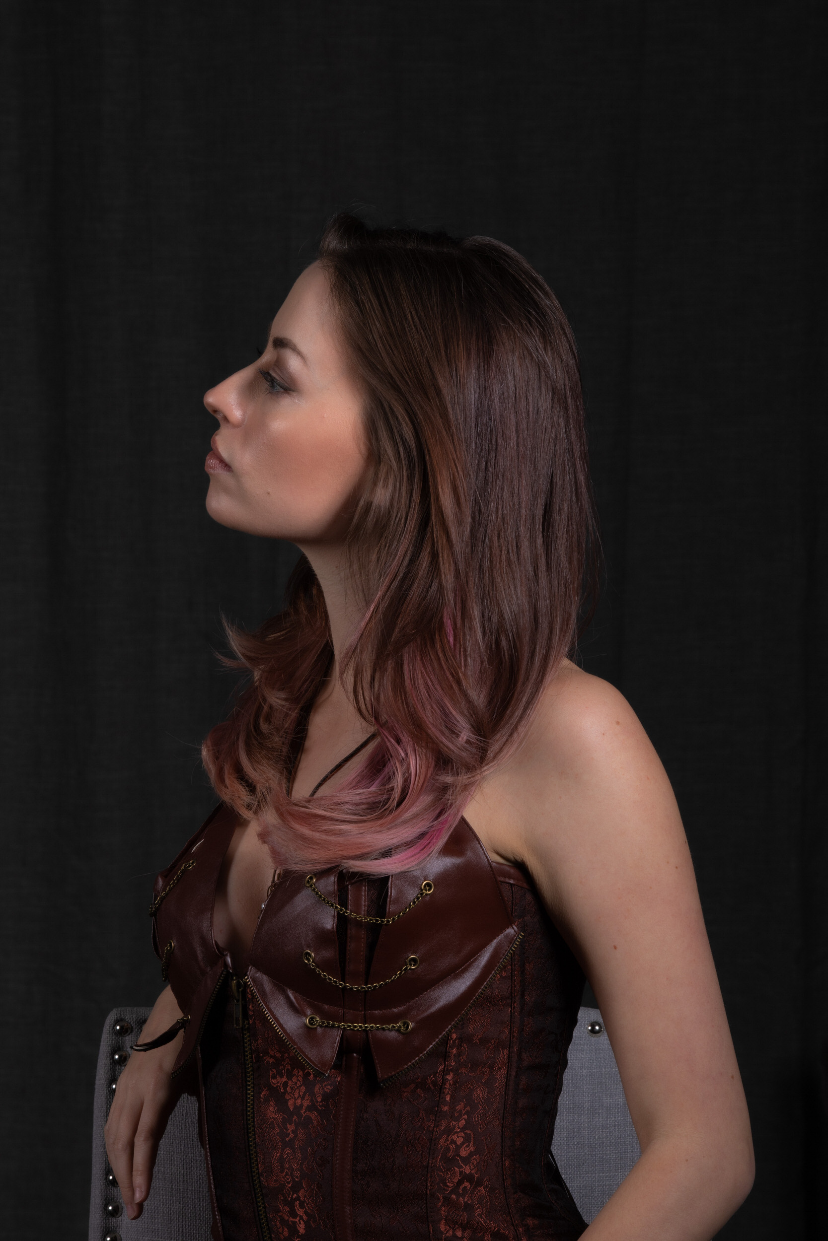 the image showcases a young woman with long, brown hair accented with pink highlights. she is positioned in profile view against a dark, almost black backdrop.  the woman wears an elaborately designed steampunkinspired corset in shades of brown and copper, featuring leather straps, buckles, and lace detailing.  the lighting is artificial, likely studio lighting, which casts soft shadows and highlights her features. the image exudes a sense of strength and individuality, emphasized by her confident posture and the striking attire.