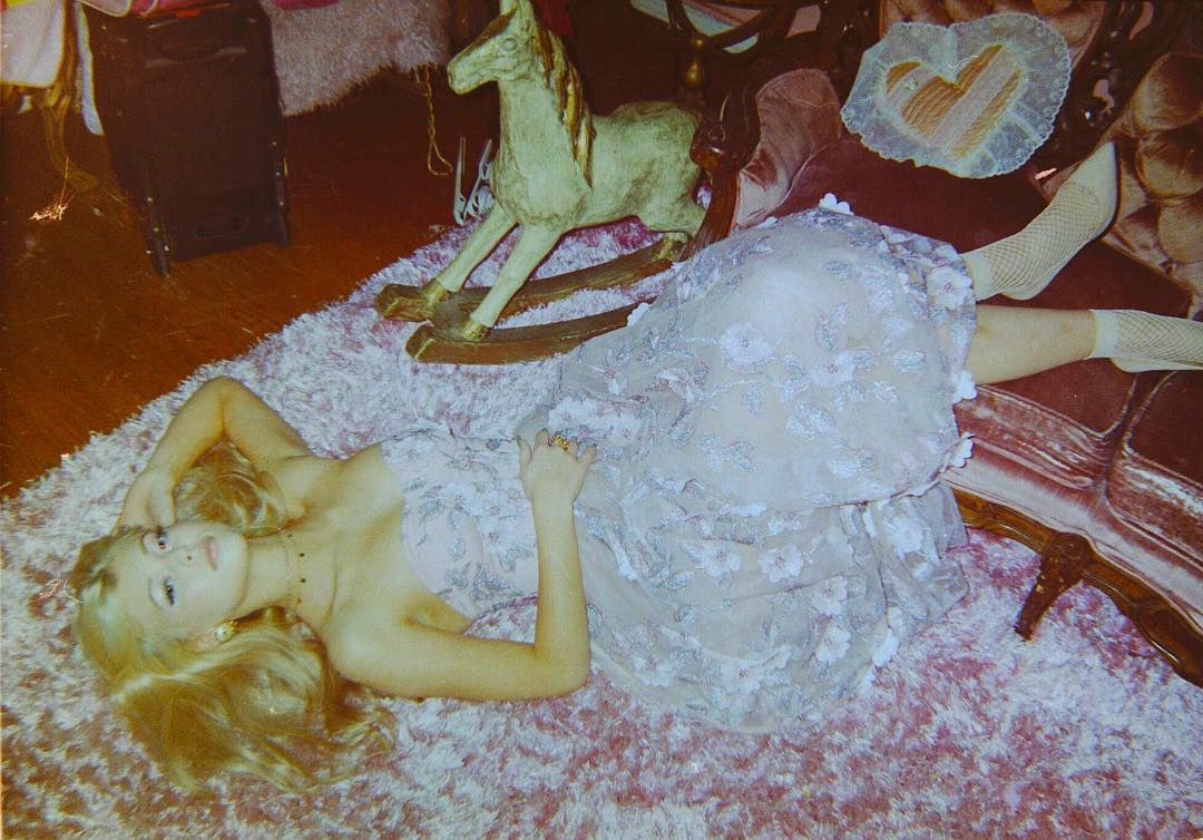 a vintagestyle photograph features a blonde woman lying on a fluffy pink carpet. she is wearing a pale pink dress with white floral embellishments and fishnet socks. next to her is a wooden rocking horse. the image also includes a piece of furniture and other decorative items, contributing to the overall vintage aesthetic.