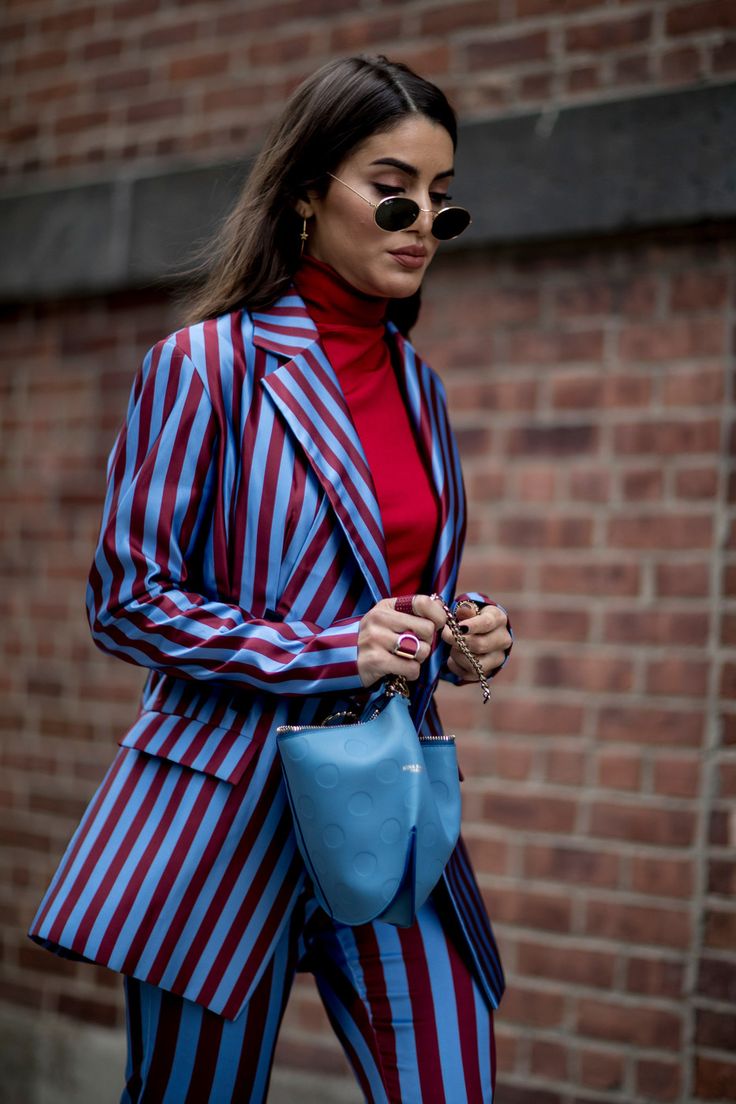 a stylish woman is captured in a street style photograph. she's wearing a vertically striped blazer and pants suit in shades of blue and maroon. underneath the blazer, she sports a vibrant red turtleneck. her accessories include roundframed sunglasses, a light blue handbag with a metallic chain strap, and several rings on her fingers. the background consists of a rustic red brick wall. the overall impression is one of sophisticated and trendy autumnal fashion.