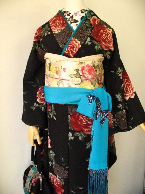 a kimono, primarily black with a floral rose pattern, is displayed on a mannequin. the kimono is cinched at the waist with a floral obi and a bright blue sash featuring butterfly designs and fringe detailing. the collar of the kimono has a contrasting pattern and a blue trim. the outfit is accessorized with a patterned bag hanging to the left.