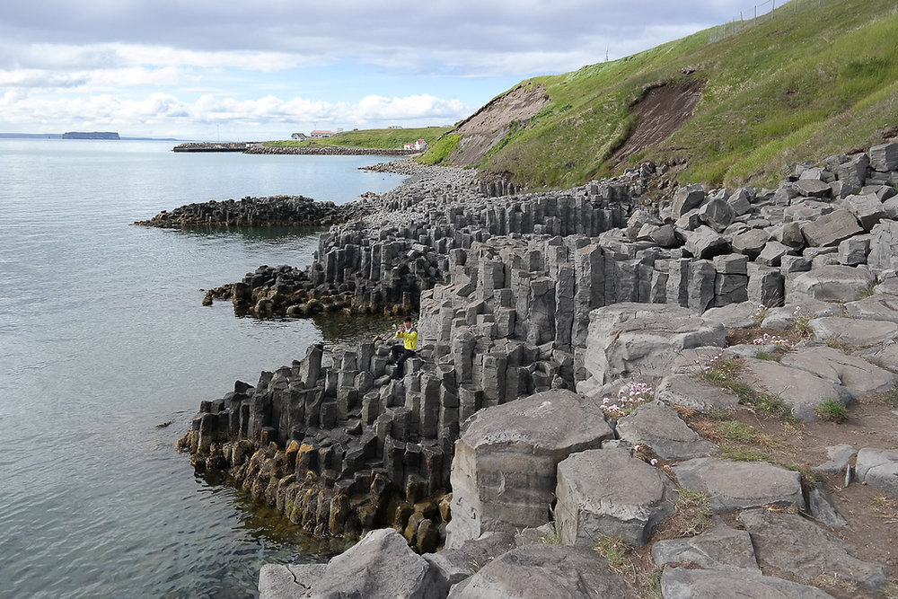 the image shows a basalt column coastline with an ocean, rocks, cliff, grass, sky, island, and harbor. there is a person wearing a yellow jacket. the basalt columns are arranged in vertical stacks, creating a unique natural formation. the sky is overcast.