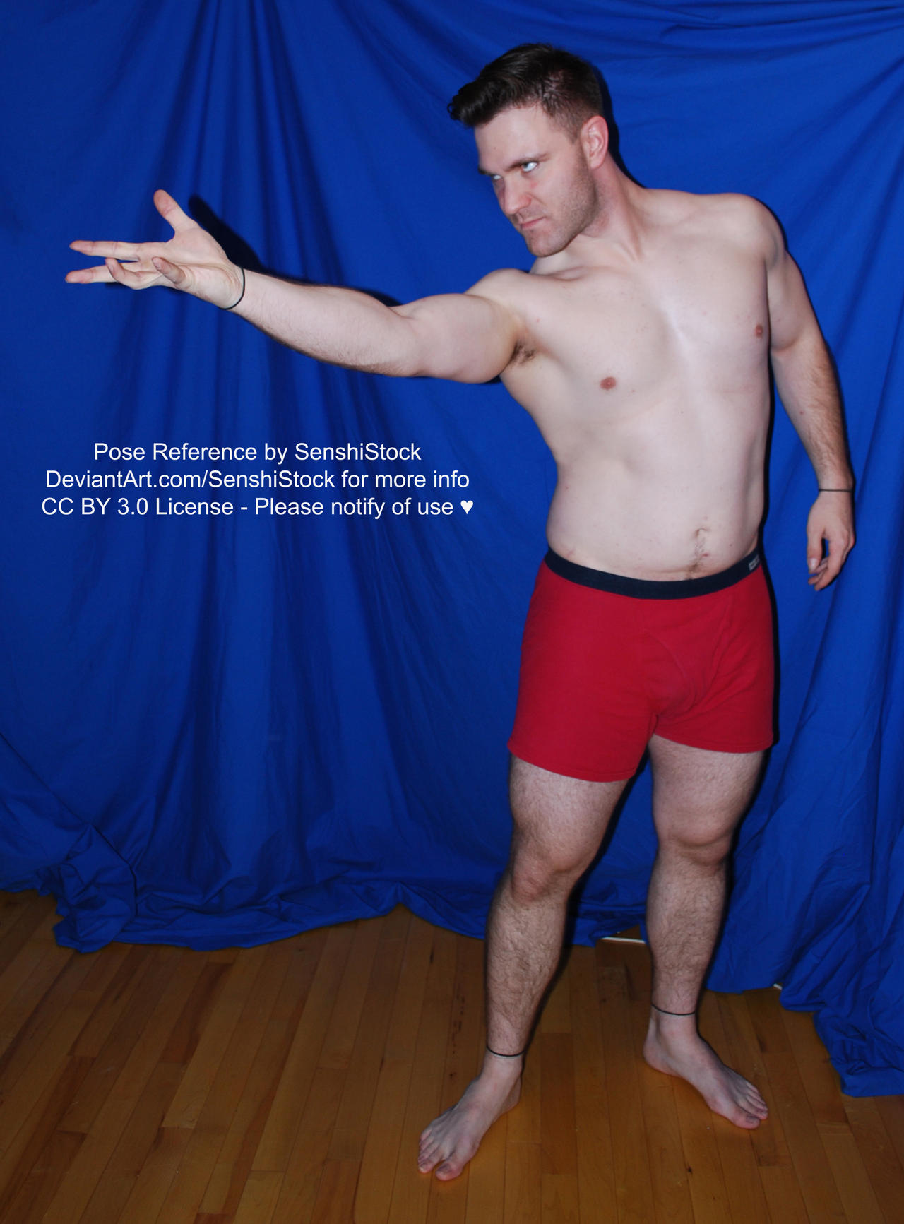 fullbody shot of a muscular male model posing against a blue backdrop.  he is shirtless, wearing red boxer shorts, and his arms are outstretched to his left. his expression is serious.  the image is presented as a pose reference for artists.  copyright information is overlaid on the image.