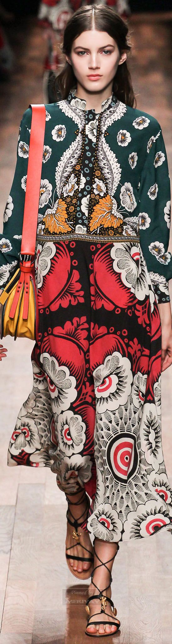 a fashion model walks down a runway wearing a vibrant, multipatterned dress. the dress features a dark green floral top and a red and black floral skirt with white accents. the model carries a yellow and red handbag and wears black lacedup sandals. the overall style is bohemian and eclectic.  the background is blurred, focusing attention on the model and her outfit.