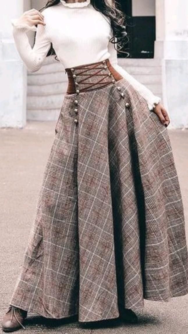 a woman stands outdoors in natural light, showcasing a stylish autumn outfit. she wears a white turtleneck sweater tucked into a highwaisted, flowing plaid skirt in shades of brown. the skirt features a brown faux leather waistband with a laceup detail and decorative buttons down the front. her shoes are not visible in the image. the photo evokes a vintage, bohemian feel with a modern twist.