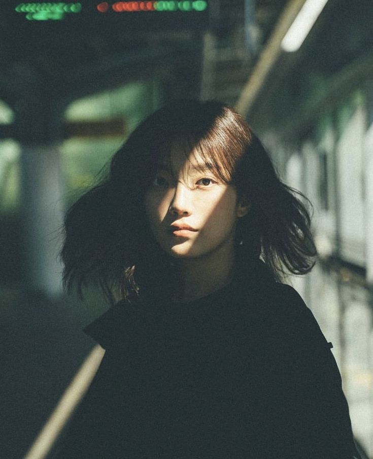 a young woman with long dark hair and a black shirt poses for a portrait while looking directly at the camera. her face is halfshadowed and halflit.  she's surrounded by what appears to be an urban environment.