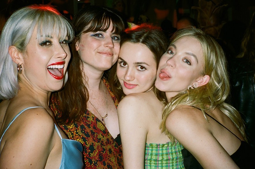 the image shows four young women huddled together in a close, candid group photo. they appear to be at a party or social gathering indoors at night.  the women are smiling and laughing, showcasing a close and happy friendship.  their attire is casual and colorful, and the photo's style suggests it was taken with film photography, giving it a vintage or retro feel. the mood is lively and joyful.