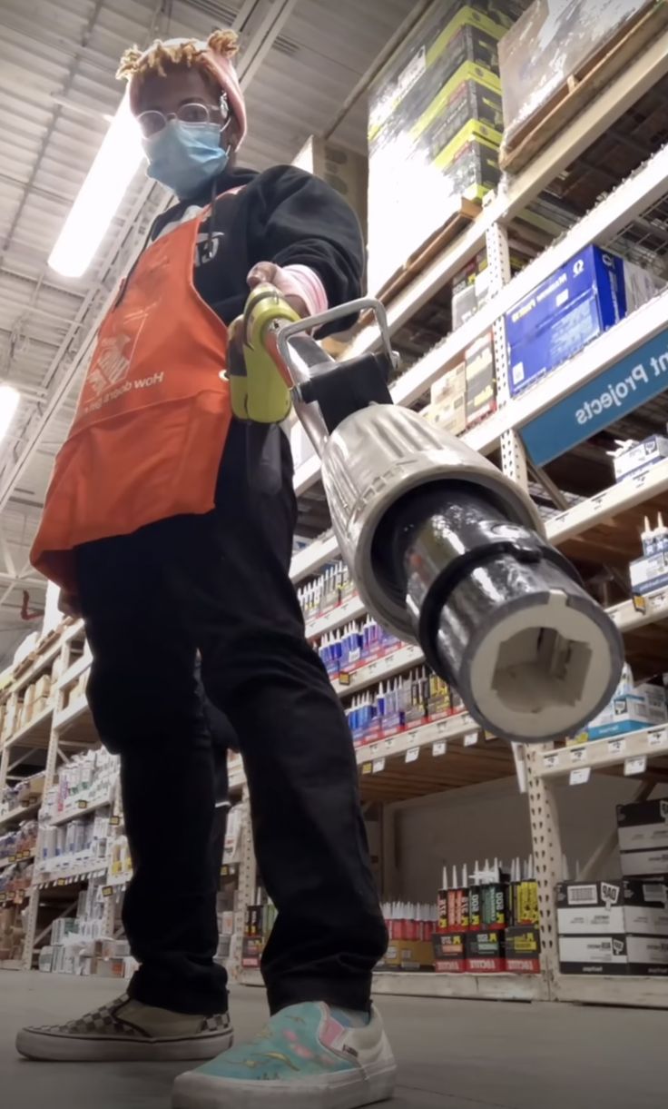 a young person wearing a home improvement store uniform, orange apron, face mask, and sunglasses, holds a tool in a home improvement store aisle.