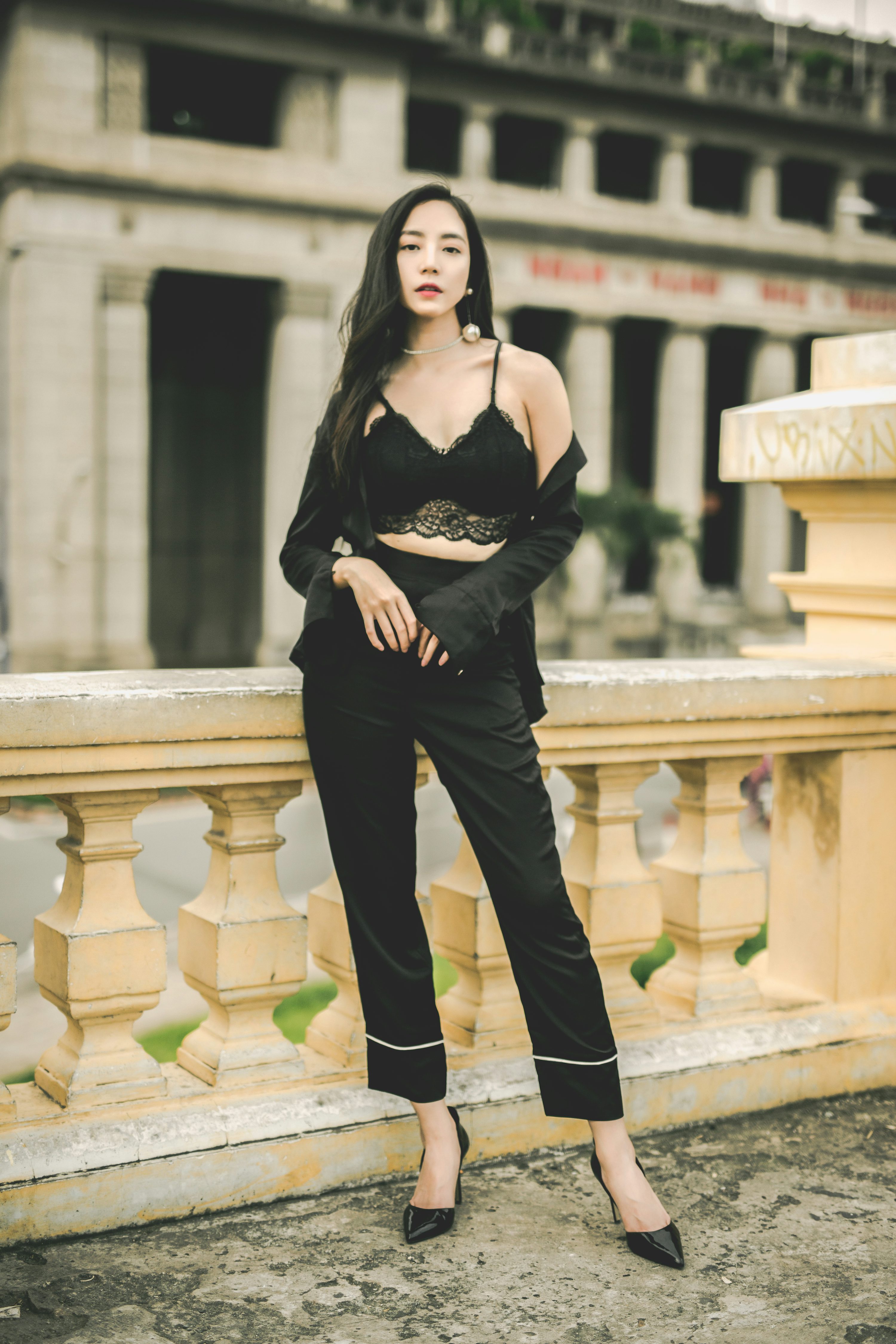 a young asian woman is standing against a beautiful vintage building. she's wearing a stylish black outfit: a lace crop top, black pants with white trim, and a black blazer. her high heels complete the elegant look. the background shows an ornate building with columns. she is looking straight at the camera with a confident expression. the image has a fashionable and sophisticated vibe.
