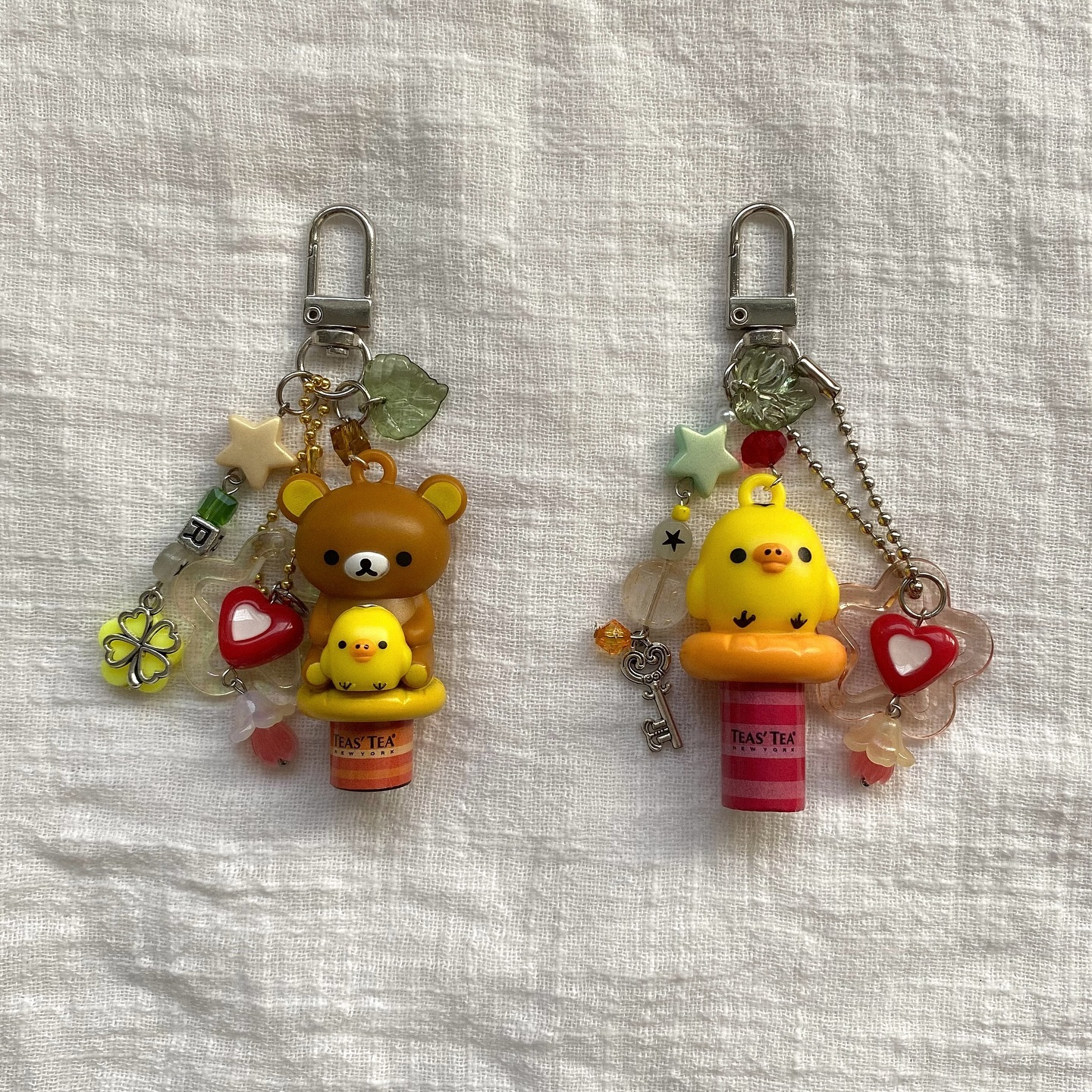 two keychains are displayed on a textured, light beige background. the keychain on the left features a brown rilakkuma bear figure holding a small chick sitting atop a miniature can of "teas' tea". various charms including a fourleaf clover, a red heart, stars, leaves, and several beads are attached around it. the second keychain showcases a yellow chick figure on a can of "teas' tea" along with similar charms such as a heart, star, leaf, beads, and a key. the metal clasps at the top of each keychain are identical. the textured background appears to be a fabric surface.