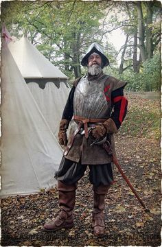 a man dressed in a medieval suit of armour, complete with helmet, stands outside a tent in a forest setting.  he holds a walking stick in his right hand and a sword at his side.  he's  looking intensely at something offcamera.