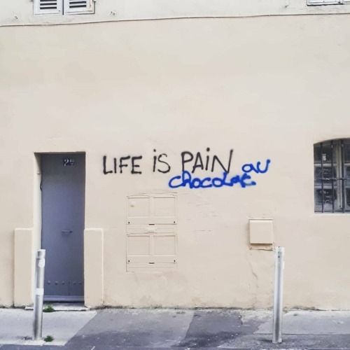 the image shows a building wall with graffiti that reads "life is pain au chocolat." there is a door on the left, a small window on the right, and a closedoff section in the middle. the writing is in black and blue spray paint. there is also a grey pole that runs from the ground up the side of the building.