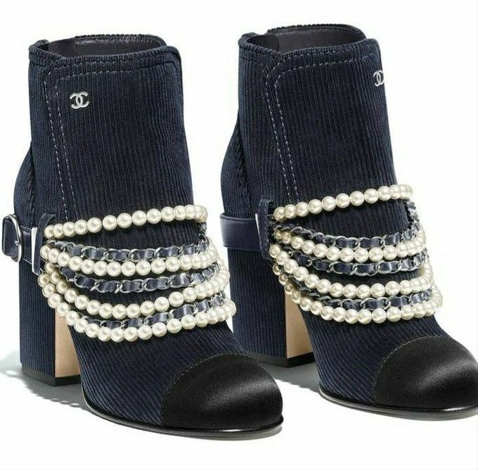 the image shows a pair of navy blue chanel ankle boots with pearl and chain embellishments. the boots have a corduroy texture on the upper part, a black satin toe cap, a chunky heel, and a buckle on one side. the boots are adorned with multiple strands of pearls and intertwined chains. the chanel logo is visible on the side of each boot.