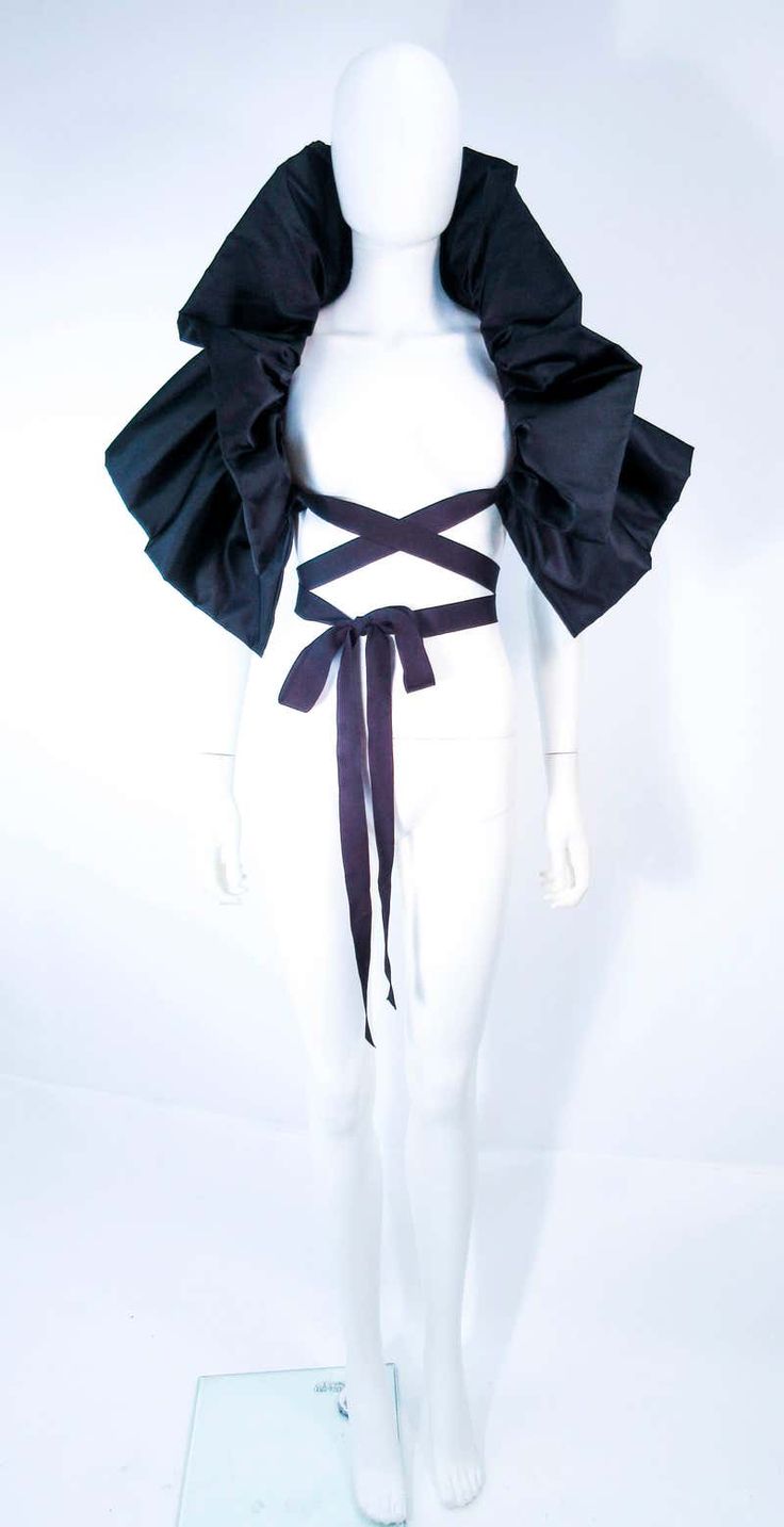 a white mannequin wearing a black ruffled collar dress with a large bow on the waist. the background is white. the dress is a simple white halter top with black ruffle detail and is cinched at the waist with a black sash that ties into a large bow.  the mannequin has no face or hands.